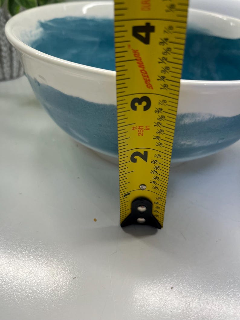 May include: White ceramic bowl with a teal blue painted band. A yellow measuring tape shows the bowl is approximately 10 cm deep. The bowl has a smooth, glossy finish and is suitable for serving food.