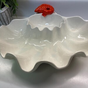 May include: A white ceramic serving dish with a wavy, shell-like design. The dish has three compartments and is topped with a bright red lobster figurine. This decorative dish is perfect for serving appetizers or snacks.