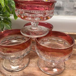 May include: A set of three clear glass dessert bowls with a pedestal base and a deep red rim. Each bowl features a textured pattern of raised circles and a band of small geometric indentations around the upper section of the glass body.