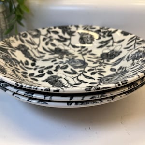 May include: A stack of three round ceramic plates featuring a black and white floral pattern. The design consists of detailed illustrations of various flowers, leaves, and vines covering the entire surface of each plate in a monochromatic style.