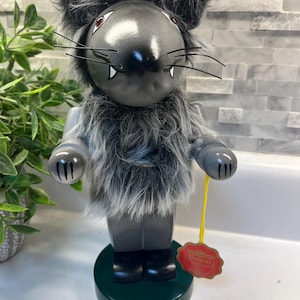 May include: A wooden nutcracker designed as a grey wolf with a smooth, rounded head, pointed ears, and long black whiskers. It features a thick, shaggy grey and white faux fur chest, grey arms with black claw details, and a red tag on a yellow string.