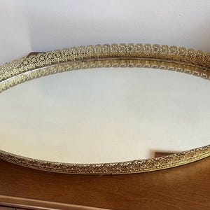 May include: An oval vanity tray featuring a reflective mirrored base and a decorative gold-toned metal rim. The raised edge of the tray displays an intricate, perforated filigree pattern that encircles the entire perimeter of the mirror.