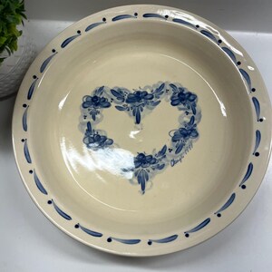 May include: A white ceramic pie plate with a blue floral design in the shape of a heart. The plate has a blue and white border with a small blue dot pattern. The text "Chuy's 1997" is written in blue on the bottom of the plate.