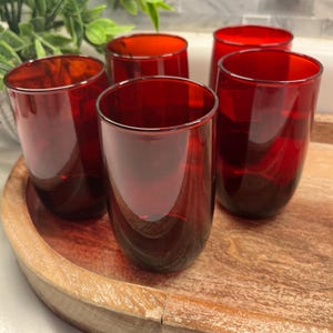 May include: Five stemless red wine glasses are arranged on a wooden serving tray. The glasses are a deep red color and have a smooth, rounded shape. The tray is light brown and has a natural wood grain.