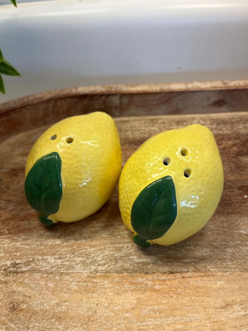 May include: A pair of lemon-shaped ceramic salt and pepper shakers. The shakers are yellow with a textured finish and green leaf details. Each shaker has three holes on top for dispensing salt or pepper.