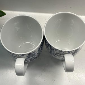 Puede incluir: Dos tazas de cer&aacute;mica blancas con un dise&ntilde;o de paisaje azul y blanco. Cada taza tiene un cuerpo redondeado, un asa blanca y una ilustraci&oacute;n detallada de edificios y &aacute;rboles. Las tazas est&aacute;n vac&iacute;as y una al lado de la otra.