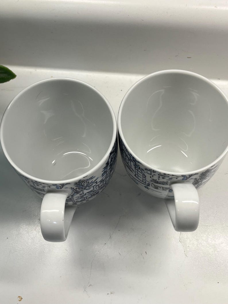 Puede incluir: Dos tazas de cer&aacute;mica blancas con un dise&ntilde;o de paisaje azul y blanco. Cada taza tiene un cuerpo redondeado, un asa blanca y una ilustraci&oacute;n detallada de edificios y &aacute;rboles. Las tazas est&aacute;n vac&iacute;as y una al lado de la otra.