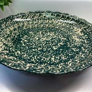 May include: A round, ceramic serving platter with a scalloped edge. The plate features a speckled pattern in dark green and cream colors. The rim has a wavy design.