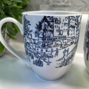 Puede incluir: Taza de cer&aacute;mica blanca con una ilustraci&oacute;n azul de una escena callejera, que incluye edificios, &aacute;rboles y personas. La taza tiene un asa curva y las palabras "IN ROUGE" son visibles. Fondo blanco.