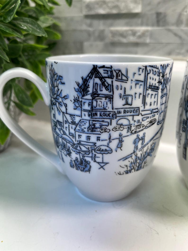 Puede incluir: Taza de cer&aacute;mica blanca con una ilustraci&oacute;n azul de una escena callejera, que incluye edificios, &aacute;rboles y personas. La taza tiene un asa curva y las palabras "IN ROUGE" son visibles. Fondo blanco.