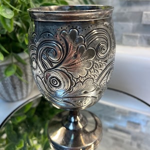 May include: A silver-toned metal goblet featuring an intricate embossed pattern of swirling scrolls, floral motifs, and textured dots. The vessel has a rounded bowl, a slender stem, and a circular base, with a polished finish and dark patina.