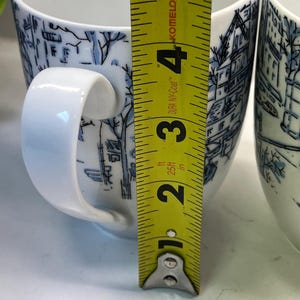 Puede incluir: Dos tazas de cer&aacute;mica blanca con ilustraciones de paisajes azules. Cada taza tiene un asa blanca y mide aproximadamente 10 cm de alto. Una cinta m&eacute;trica amarilla se coloca junto a las tazas.