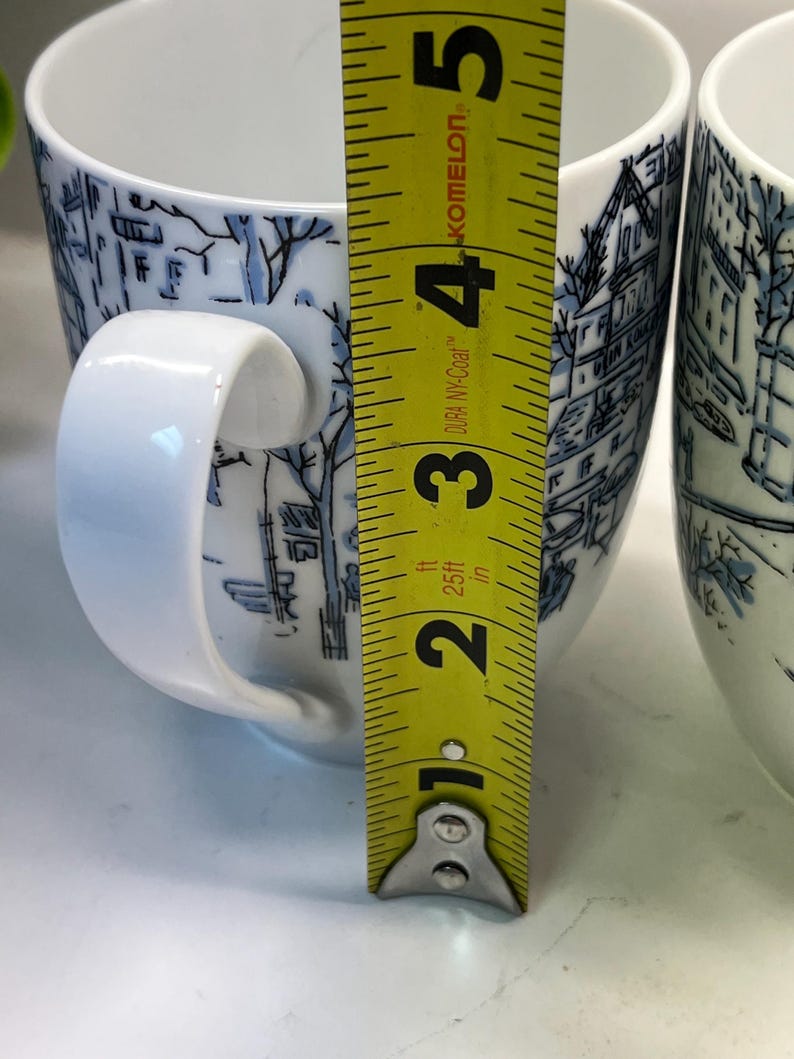Puede incluir: Dos tazas de cer&aacute;mica blanca con ilustraciones de paisajes azules. Cada taza tiene un asa blanca y mide aproximadamente 10 cm de alto. Una cinta m&eacute;trica amarilla se coloca junto a las tazas.