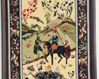 May include: Framed tapestry featuring a pastoral scene with figures on horseback and in traditional attire. The artwork has a cream-colored background with a border of floral and animal motifs in shades of brown, green, and blue. The frame is dark brown.
