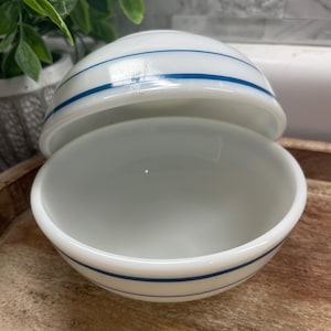 May include: A white, vintage-style bowl with two thin, horizontal blue stripes. The bowl has a matching lid, and both pieces are made of a glossy, opaque material. The bowl is open, revealing its interior.