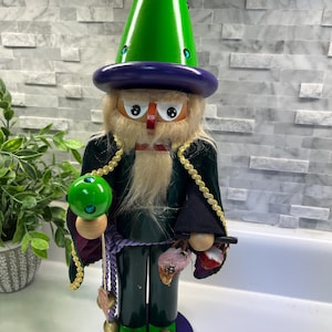 May include: A wooden wizard nutcracker with a green pointed hat, purple hat brim, and a long white beard. The wizard wears a green velvet coat with gold trim and holds a green ball. The base is purple with the text "The Wizard of Oz".