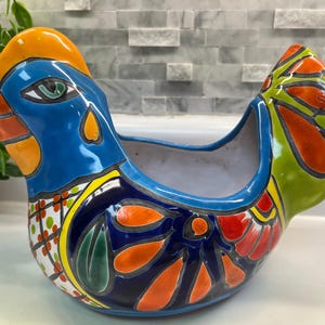 May include: A vibrant, hand-painted ceramic chicken planter. The planter features a blue body, orange comb, and green tail feathers, with colorful floral and leaf designs. This decorative piece is ideal for small plants or as a kitchen accent.