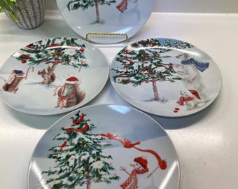 May include: Set of four white ceramic plates with winter holiday scenes. Each plate features a snowy landscape with a decorated Christmas tree and a snowman, squirrel, or polar bear. Red ribbons and accents add color.