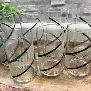 May include: A set of seven clear drinking glasses with a green diagonal stripe design. The glasses are cylindrical with a vertical striped pattern. They are arranged on a wooden tray.