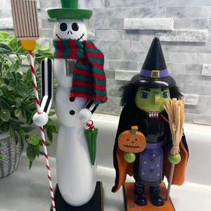 May include: Two wooden nutcracker figurines. The left figure is a white snowman with a green top hat, a red and green striped scarf, and a striped staff. The right figure is a green-faced witch in a black hat and cape, holding a pumpkin.