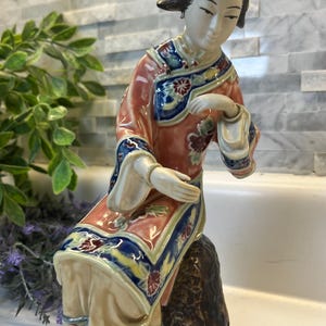 May include: A glazed porcelain figurine of a person with dark hair styled in an updo with floral accents, wearing a coral, blue, and cream-colored robe with intricate floral patterns, seated on a textured brown rock-like base with a smooth finish.