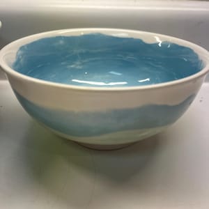 May include: A white ceramic bowl with a light blue interior and a blue band around the exterior. The bowl has a simple, elegant design, suitable for serving or display.