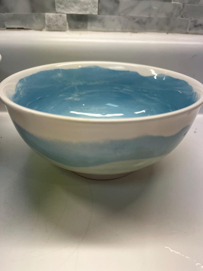 May include: A white ceramic bowl with a light blue interior and a blue band around the exterior. The bowl has a simple, elegant design, suitable for serving or display.