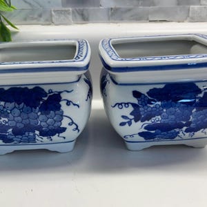 May include: Two square ceramic planters featuring a white base with blue hand-painted grape vine patterns. Each planter has a flared rim with a blue border and sits on four small feet, showcasing a classic blue and white color scheme.
