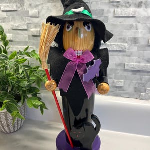 May include: A Halloween-themed wooden nutcracker in the shape of a witch. The witch wears a black hat with a green band, a black outfit, and holds a broom. A black cat sits on the purple base. The base has the number "2005" on it.