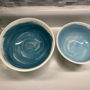 May include: Two ceramic bowls with white rims. The interior of the larger bowl is painted with a dark teal swirl design, while the smaller bowl has a light blue swirl design. The bowls are set against a neutral background.