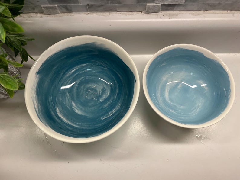 May include: Two ceramic bowls with white rims. The interior of the larger bowl is painted with a dark teal swirl design, while the smaller bowl has a light blue swirl design. The bowls are set against a neutral background.