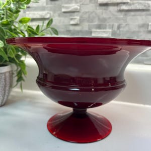May include: A deep red glass urn with a wide, flared rim and a pedestal base. The urn has a smooth, glossy finish and a classic, elegant shape. The decorative piece is likely used for display or as a decorative vase.