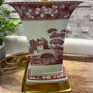 May include: A vintage table lamp with a burgundy and white floral and landscape design. The lamp has a gold-colored base and a matching top. The lamp's shade features a detailed illustration of a bridge and trees.