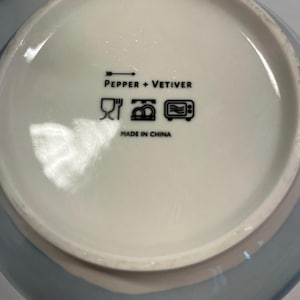 May include: A ceramic bowl with a light blue exterior and a white interior. The bowl's interior features the text "PEPPER + VETIVER" and symbols indicating it is dishwasher and microwave safe. The bowl is made in China.