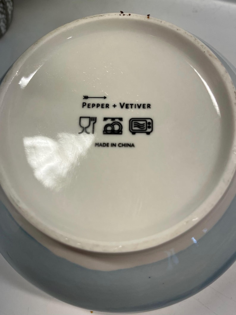 May include: A ceramic bowl with a light blue exterior and a white interior. The bowl's interior features the text "PEPPER + VETIVER" and symbols indicating it is dishwasher and microwave safe. The bowl is made in China.