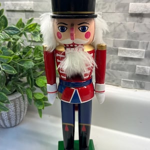 May include: A wooden nutcracker figurine, featuring a black top hat with gold trim and a red flame detail. The nutcracker has a red jacket, blue pants, and a white beard. It stands on a green base, a classic holiday decoration.