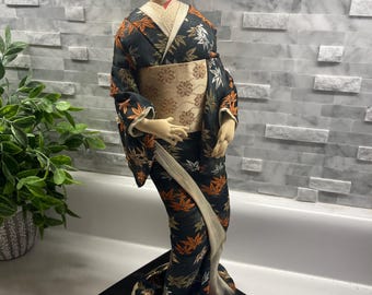 Puede incluir: Una muñeca japonesa geisha, de pie sobre una base cuadrada negra. La muñeca lleva un kimono negro con estampados de hojas doradas y naranjas, un obi beige y un peinado negro tradicional. La cara de la muñeca está pintada con rasgos delicados.