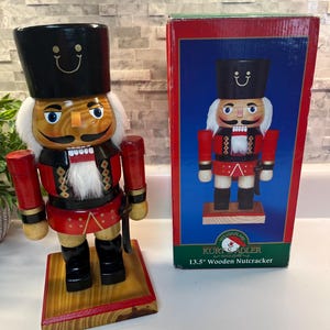 May include: A 13.5-inch wooden nutcracker figure wearing a black hat, red jacket, and black boots, standing on a wooden base. Beside it is a rectangular cardboard box featuring an image of the same nutcracker and the text Kurt S. Adler.