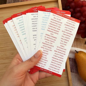 May include: A hand holds a stack of red-bordered stickers with a white background. The stickers list various holidays and observances, such as New Year's Day, Valentine's Day, and Christmas Day. The stickers are from the Cerise Sticker Shop.