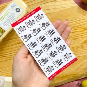 May include: A sheet of white sticker paper with 12 black and white stickers that say "get nails done" with a nail polish bottle icon. The stickers are arranged in a grid pattern. The sticker sheet is on a wooden surface.