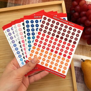 May include: A hand holds several sheets of numbered circle stickers in various colors, including red, orange, blue, and pink. The stickers are arranged in rows and columns, with numbers 1 through 31. The text "CERISE STICKER SHOP" is printed on the top of the sheets.