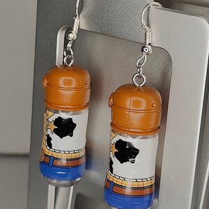 May include: A pair of silver dangle earrings featuring a brown plastic bottle with a white and brown cartoon image of Woody from Toy Story. The bottle has a blue bottom and a silver hook.