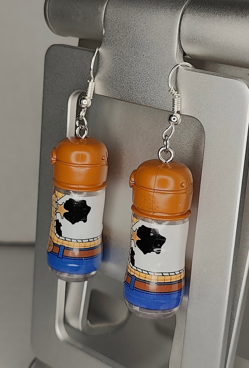 May include: A pair of silver dangle earrings featuring a brown plastic bottle with a white and brown cartoon image of Woody from Toy Story. The bottle has a blue bottom and a silver hook.
