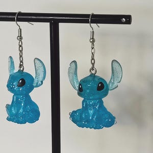 Pu&ograve; includere: Un paio di orecchini Stitch blu glitterati. Ogni orecchino presenta una piccola figura di Stitch seduta con grandi orecchie e occhi neri, sospesa da una catena e un gancio color argento. Gli orecchini sono esposti su un supporto nero.