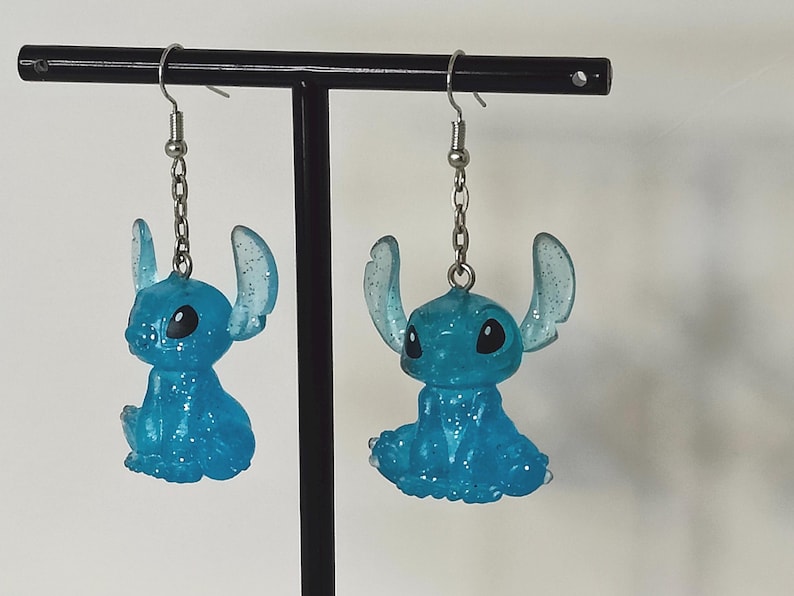 Pu&ograve; includere: Un paio di orecchini Stitch blu glitterati. Ogni orecchino presenta una piccola figura di Stitch seduta con grandi orecchie e occhi neri, sospesa da una catena e un gancio color argento. Gli orecchini sono esposti su un supporto nero.