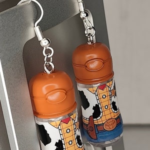 May include: A pair of dangle earrings featuring clear plastic bottles with a printed image of Woody from Toy Story. The bottles are topped with a brown cap and the earrings have silver hooks.