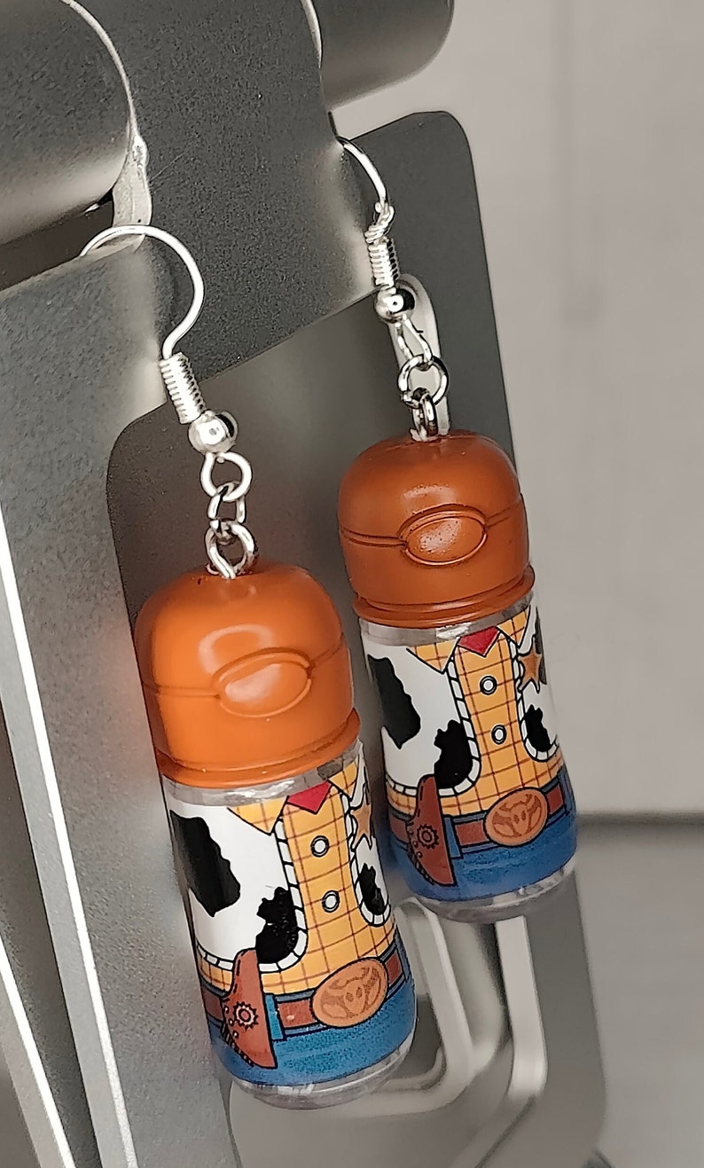 May include: A pair of dangle earrings featuring clear plastic bottles with a printed image of Woody from Toy Story. The bottles are topped with a brown cap and the earrings have silver hooks.