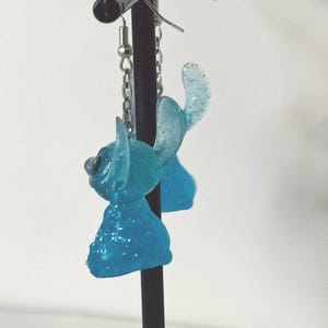 Pu&ograve; includere: Un paio di orecchini in resina blu chiaro e glitterata. Ogni orecchino presenta un design stilizzato del personaggio con grandi orecchie e una testa piccola. Gli orecchini pendono da ganci e catenelle color argento.