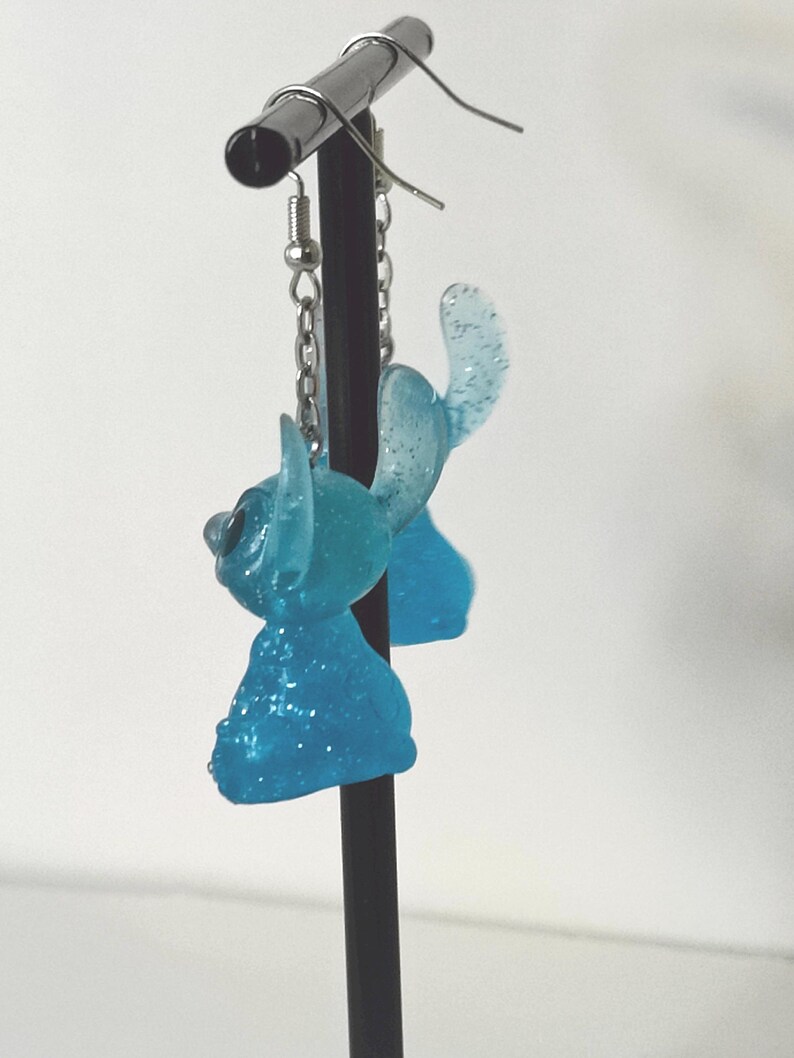 Pu&ograve; includere: Un paio di orecchini in resina blu chiaro e glitterata. Ogni orecchino presenta un design stilizzato del personaggio con grandi orecchie e una testa piccola. Gli orecchini pendono da ganci e catenelle color argento.