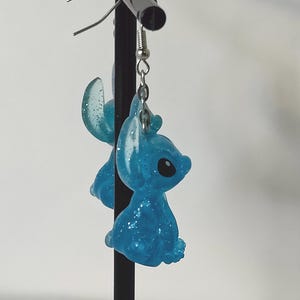 Pu&ograve; includere: Un paio di orecchini in resina glitterata blu con un design a personaggio dei cartoni animati. Gli orecchini hanno un gancio color argento e sono sospesi da un espositore in metallo nero. Il personaggio ha grandi occhi neri e una testa piccola.